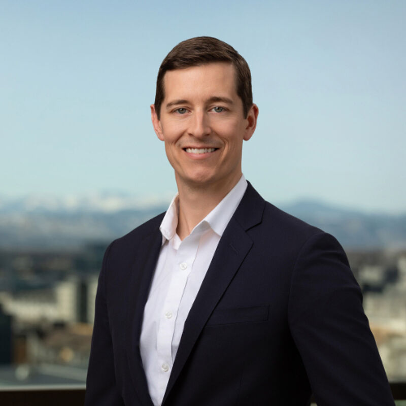 The image shows a man in a suit and white shirt, smiling confidently at the camera. He has short brown hair and fair skin. The background features a blurred cityscape with a blue sky. The overall impression is professional and approachable.